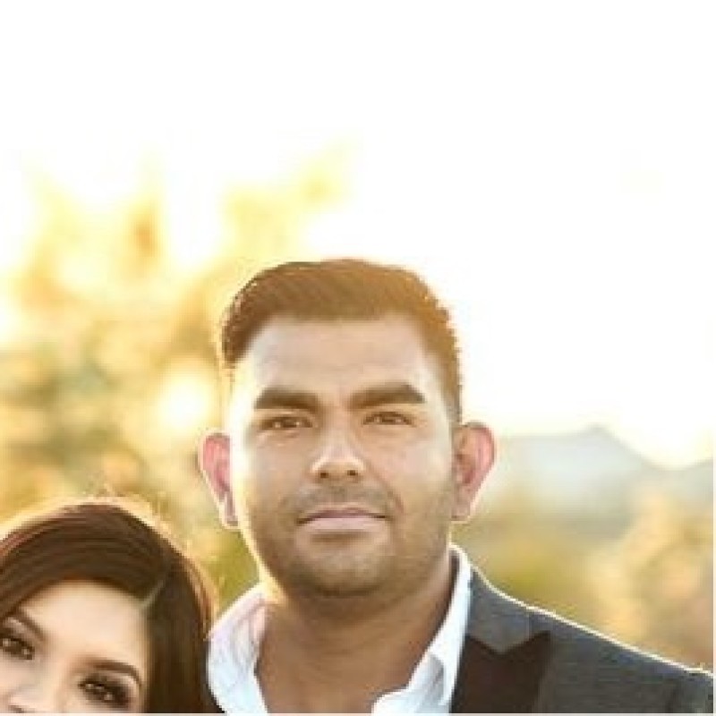 A man in a suit jacket and white shirt stands outdoors in sunlight, with part of another person's head visible beside him, possibly near a Menort Landscaping project.