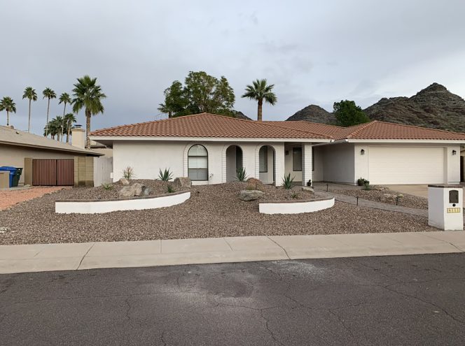 Single-story house with white exterior, red-tiled roof, arched windows, desert landscaping with gravel and rocks, and palm trees in the background.