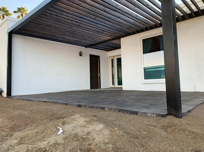 Covered patio with black tiled flooring and pergola attached to a white building; adjacent area is bare dirt without landscaping.