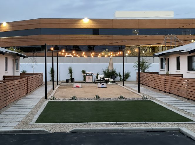 A backyard area with string lights, a barbecue grill, patio seating, and a rectangular gravel section bordered by grass and wooden fences, with buildings in the background.