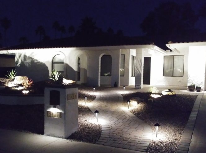A single-story white house with exterior pathway lights illuminating a stone walkway and desert landscaping at night.