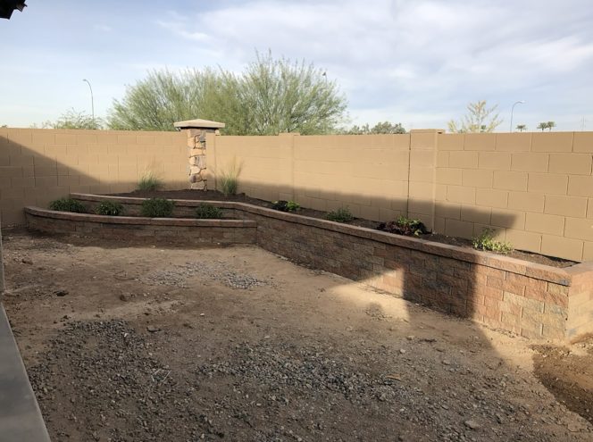 A backyard with unfinished ground, tiered brick planters along a tan block wall, small green plants, and a stone pillar in the corner.